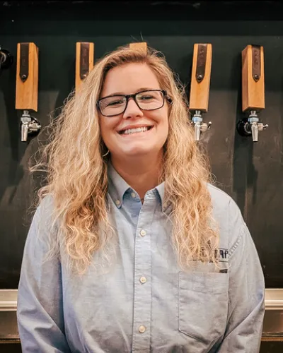 Blonde woman in a button down shirt with glasses smiling and standing in front of a row of taps.