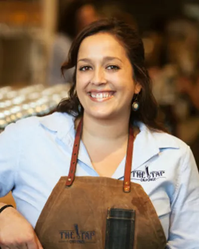 Brunette woman leaning against a bar, wearing a button down and an apron, smiling.